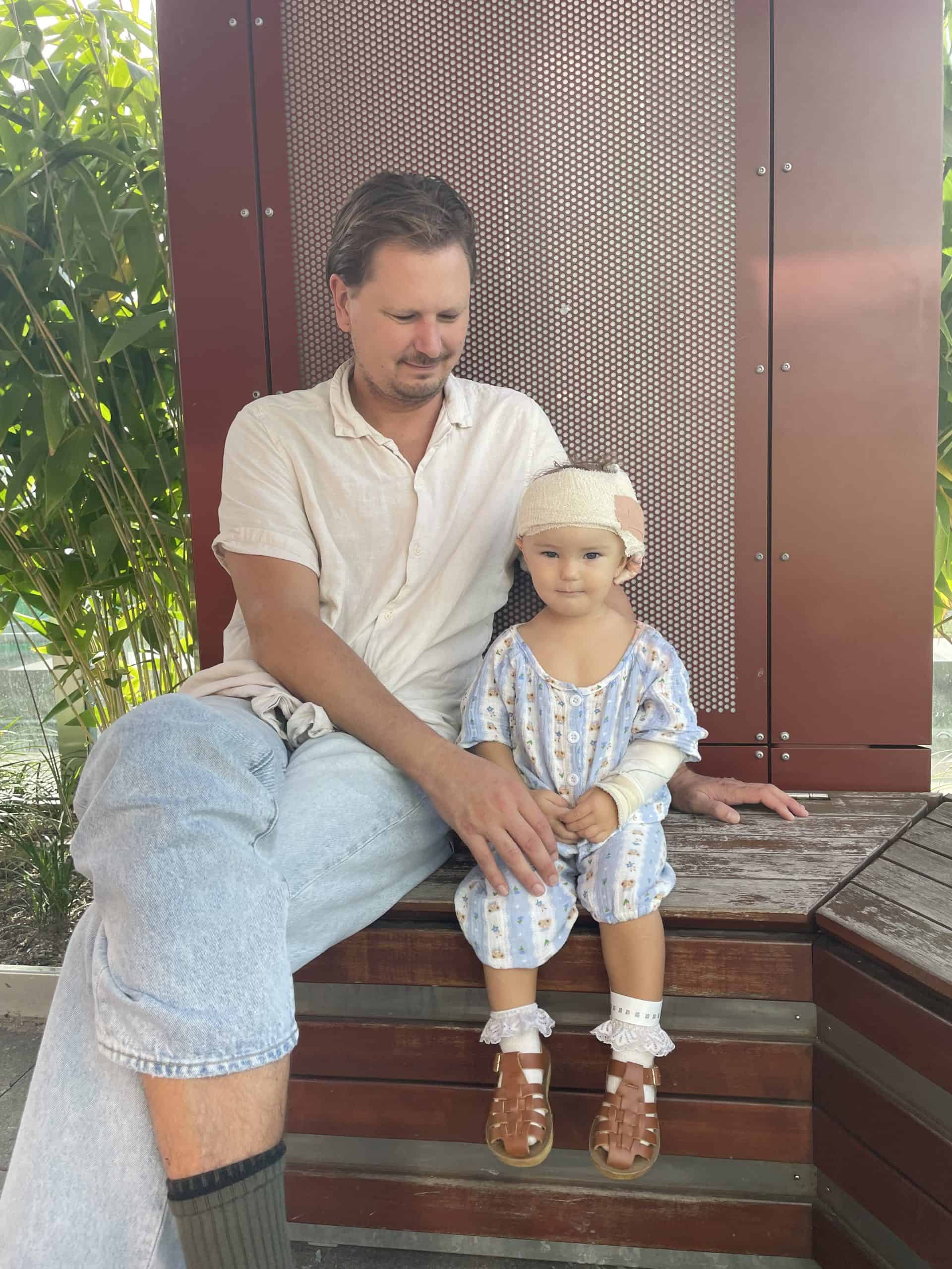 Girl with sagittal synostosis sitting with her father outdoors, wearing a bandage on her head following craniofacial surgery
