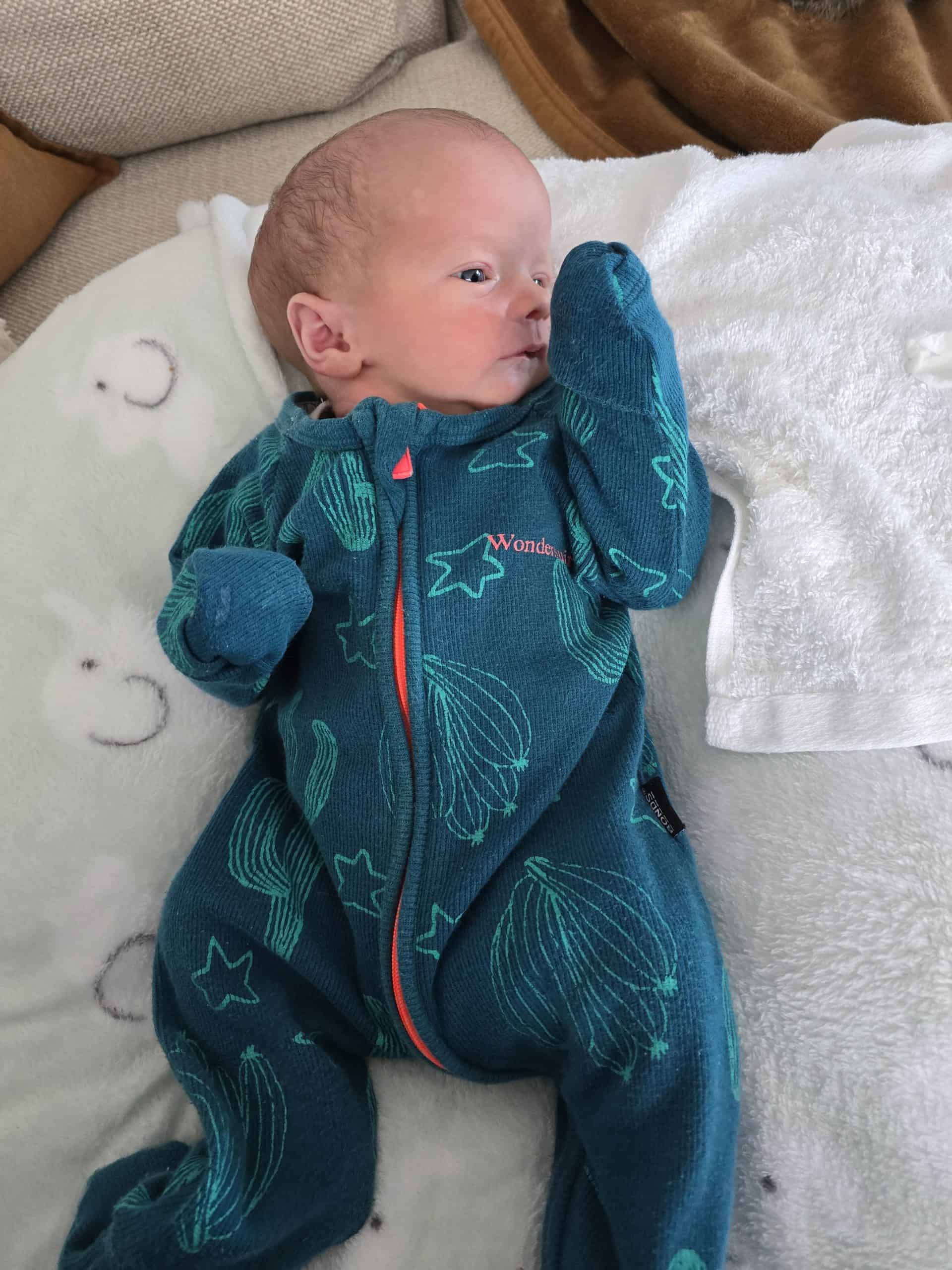 Infant baby in blue onesie lying on a bed with white blanket, facing right.