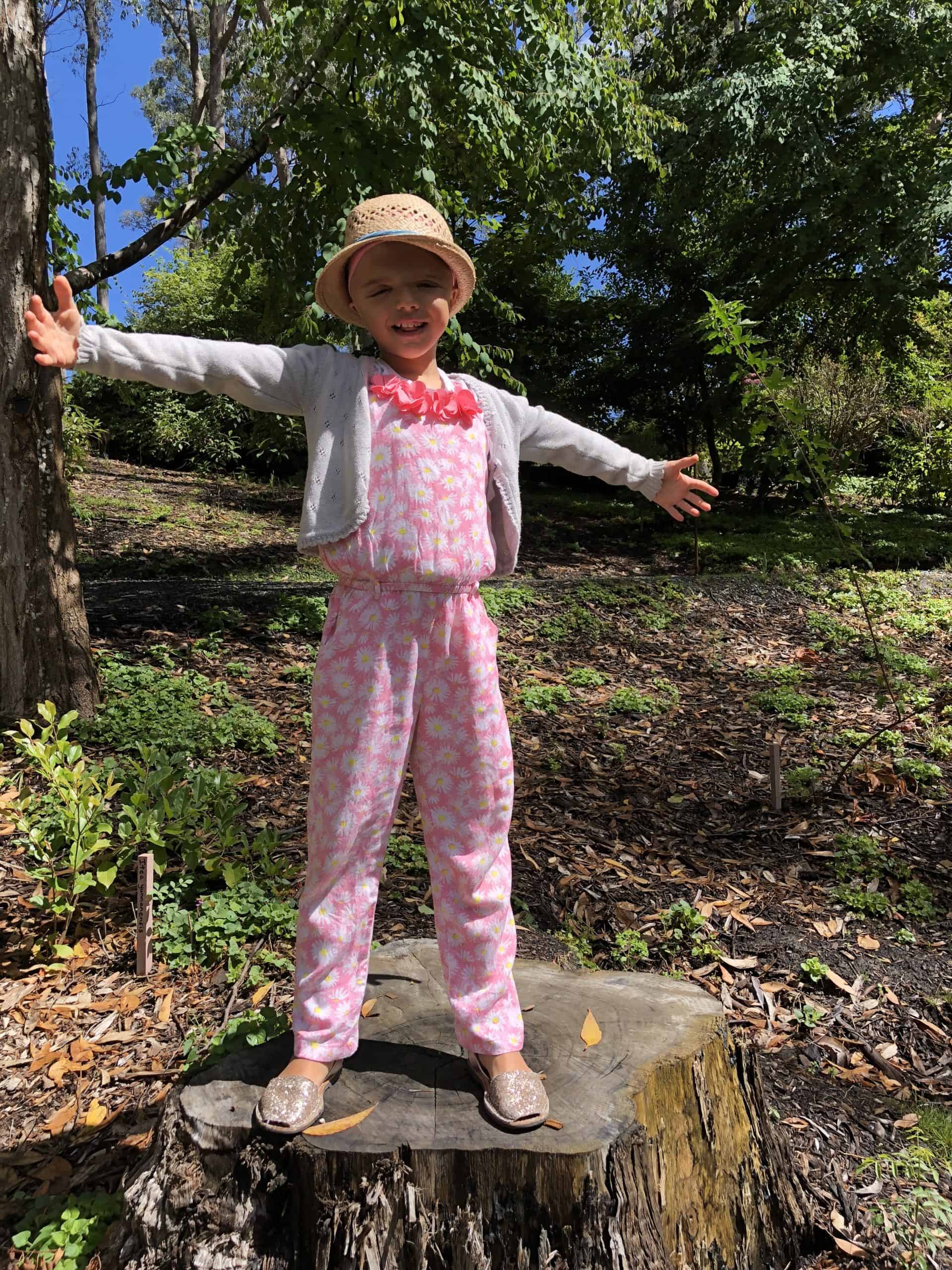Girl with Saethre-Chotzen syndrome standing on a tree stump with arms outstretched outdoors.