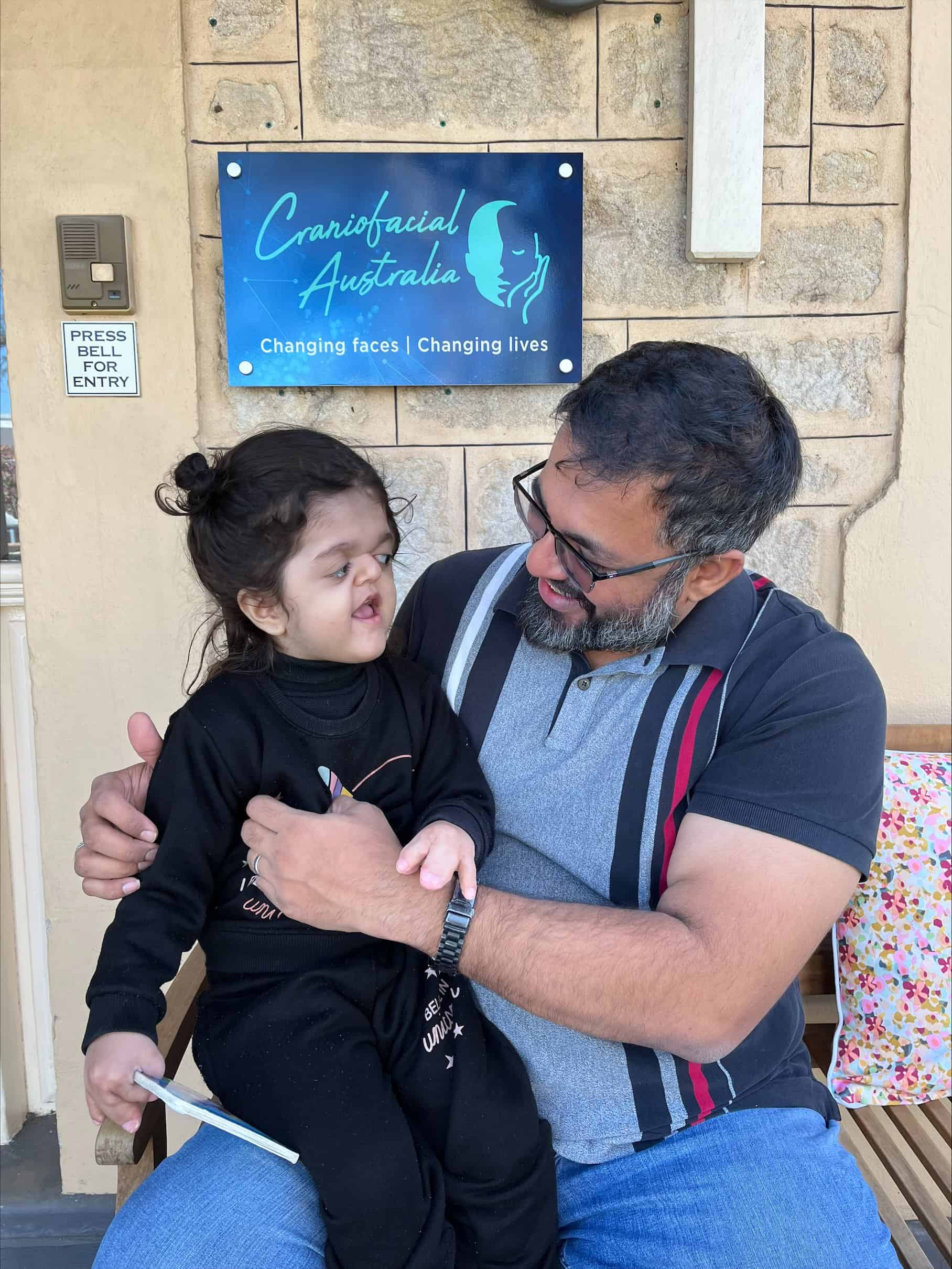 Father from Pakistan holding daughter who has Apert syndrome, sitting in front of the Craniofacial Australia office in North Adelaide