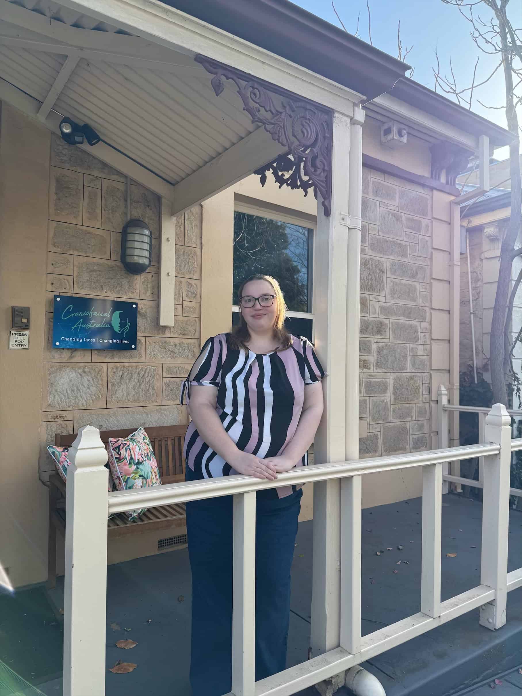 Comfortable woman standing on porch of craniofacial clinic in Australia, smiling for photo.