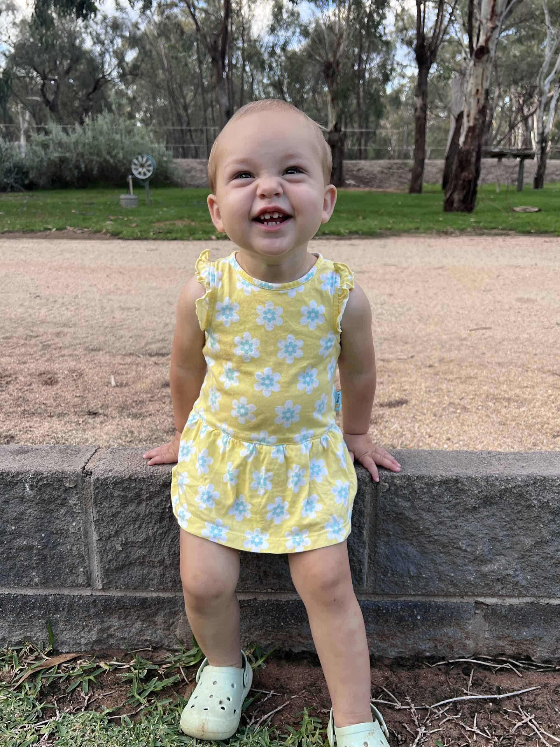 A smiling young girl in a yellow floral dress sitting on a low stone wall outdoors.