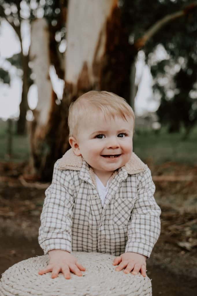 Smiling toddler outdoors near a tree in a checkered shirt.