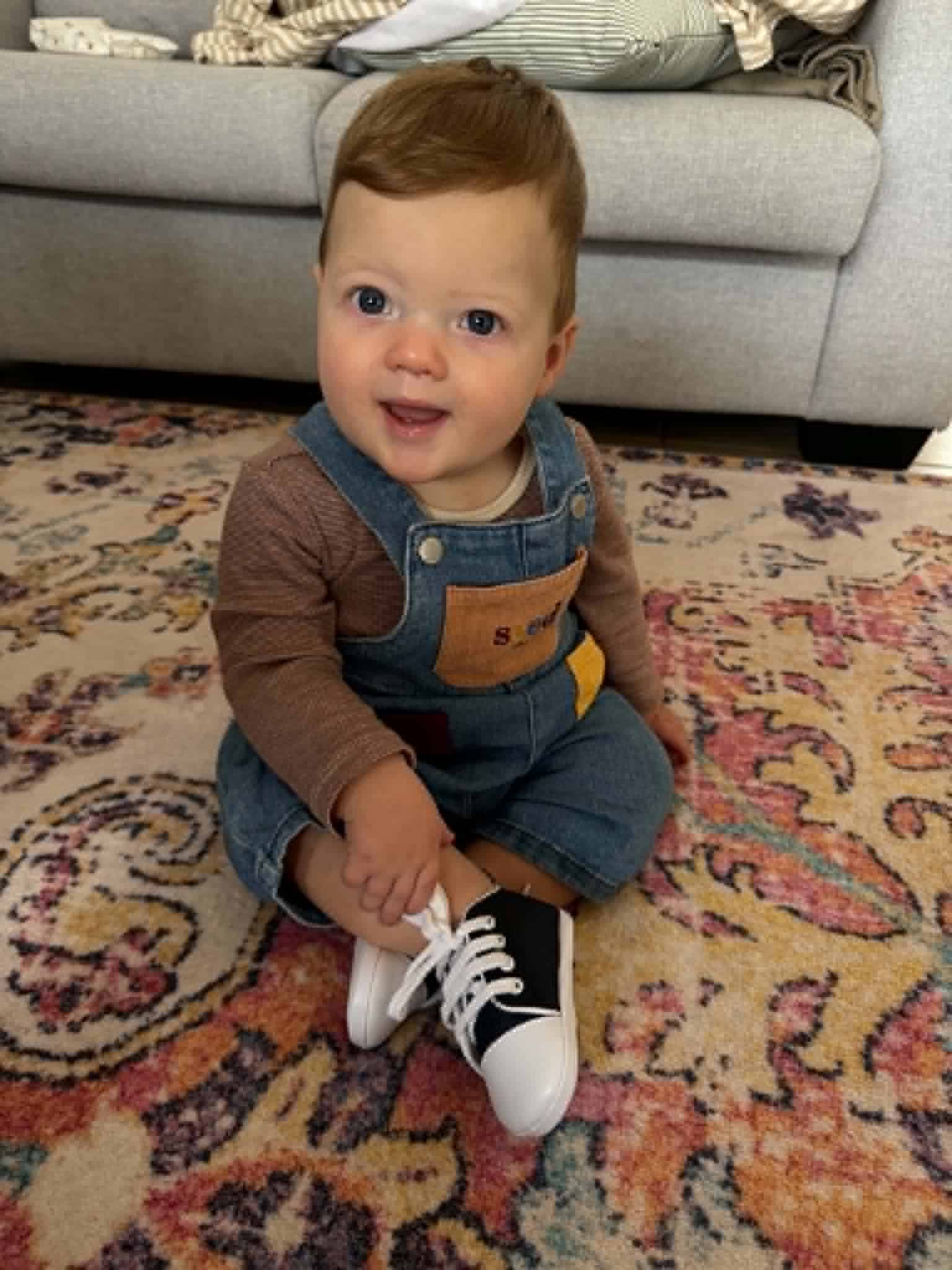 Adorable toddler sitting on a colourful rug wearing denim overalls and sneakers.