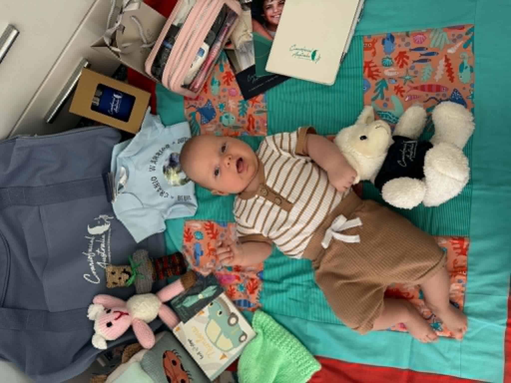 Baby lying on a bed surrounded by toys and books, with medical supplies nearby.