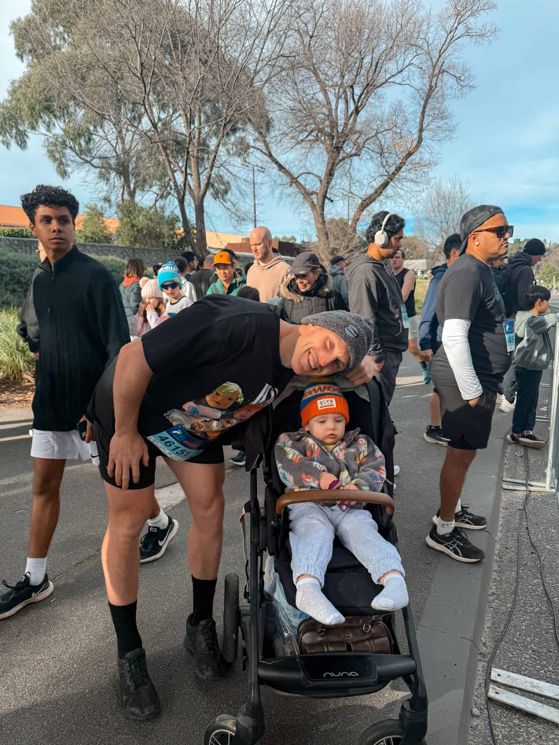 Runner with a stroller and child at a race event in a park with trees and participants.