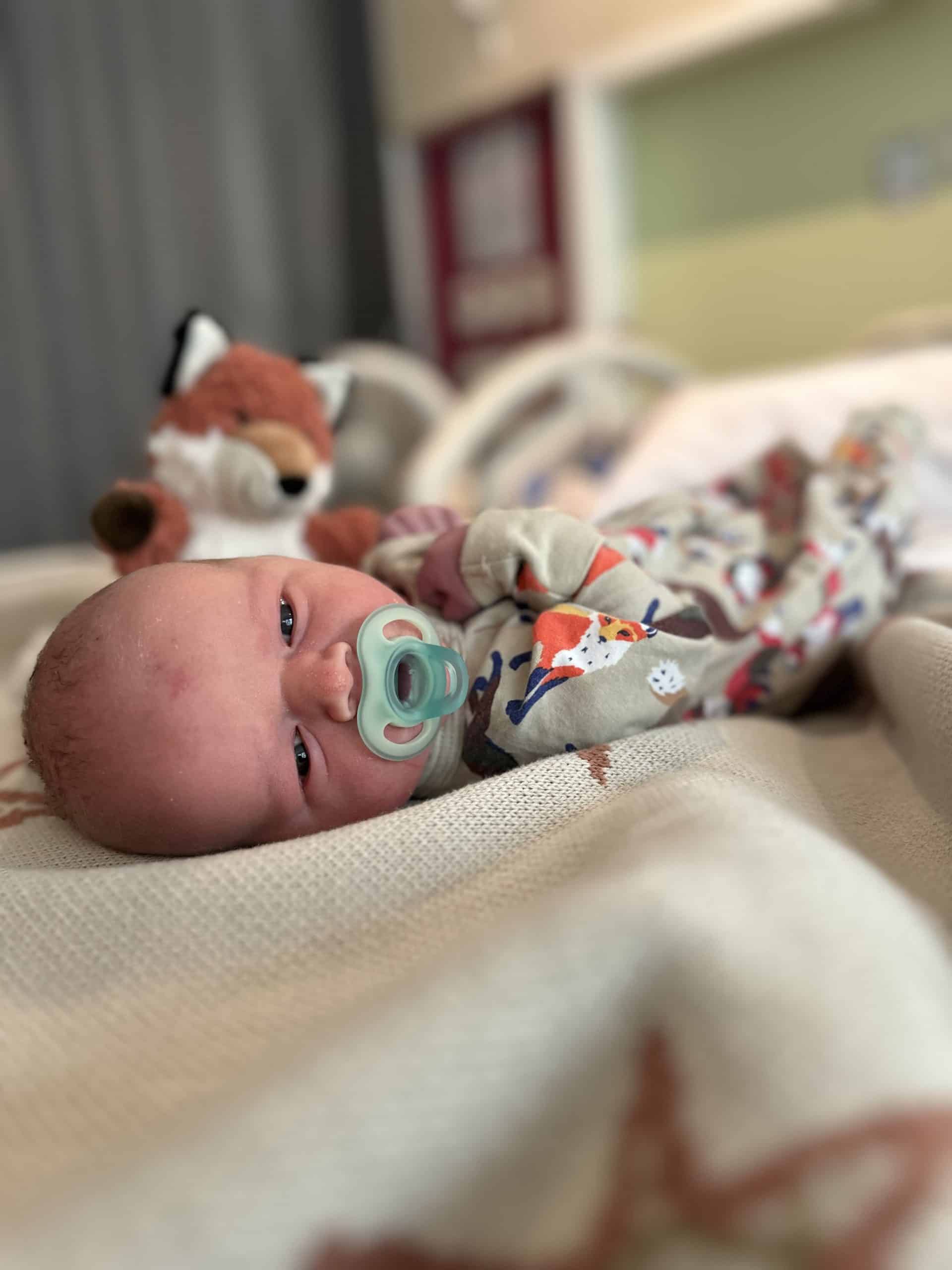 Infant with a pacifier lying in hospital bed with teddy bear in background.