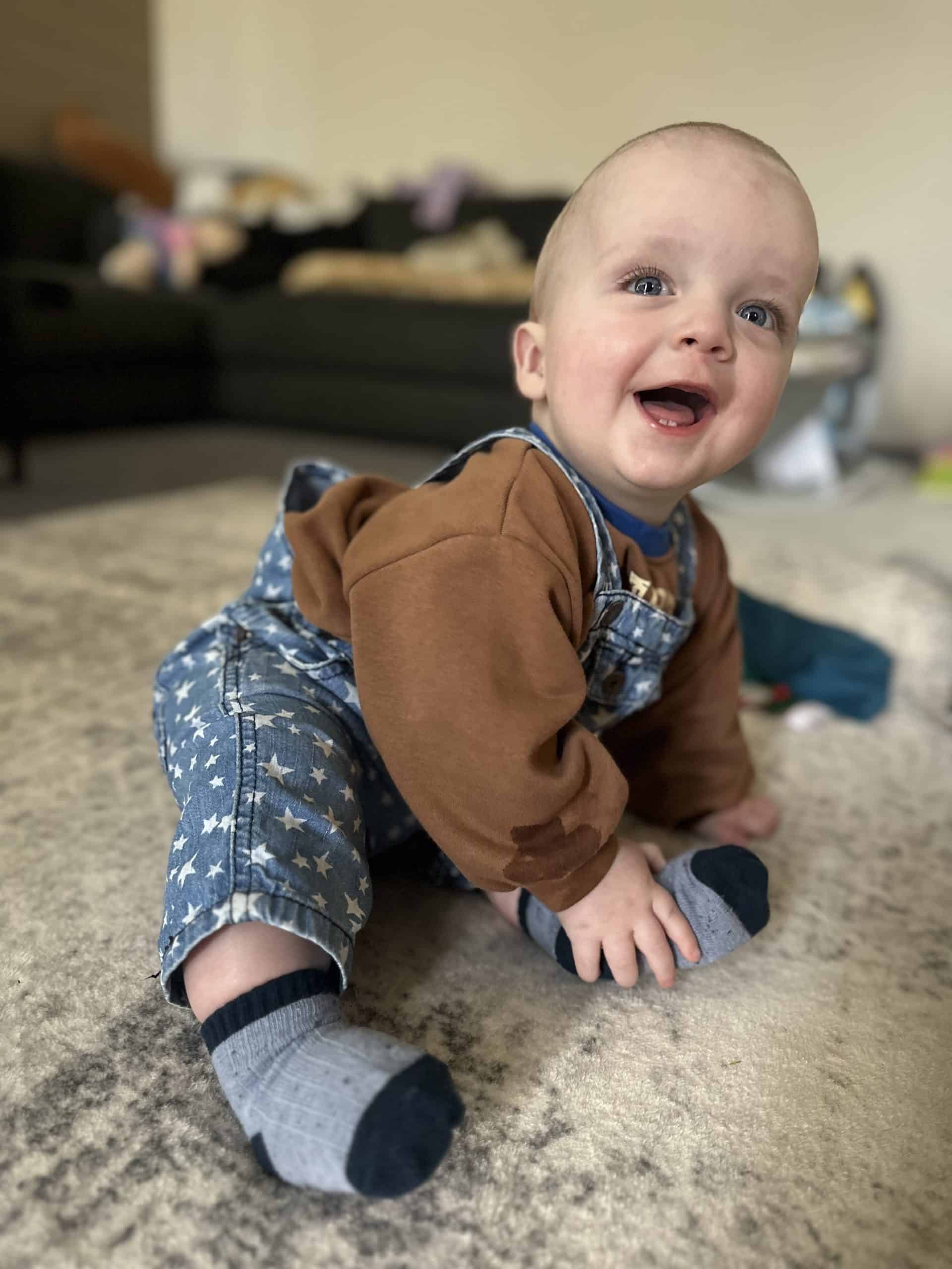 Smiling baby sitting on carpet in a cozy living room with a sofa in the background.