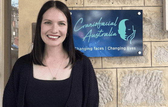 Smiling woman standing outside Craniofacial Australia clinic with logo in background.