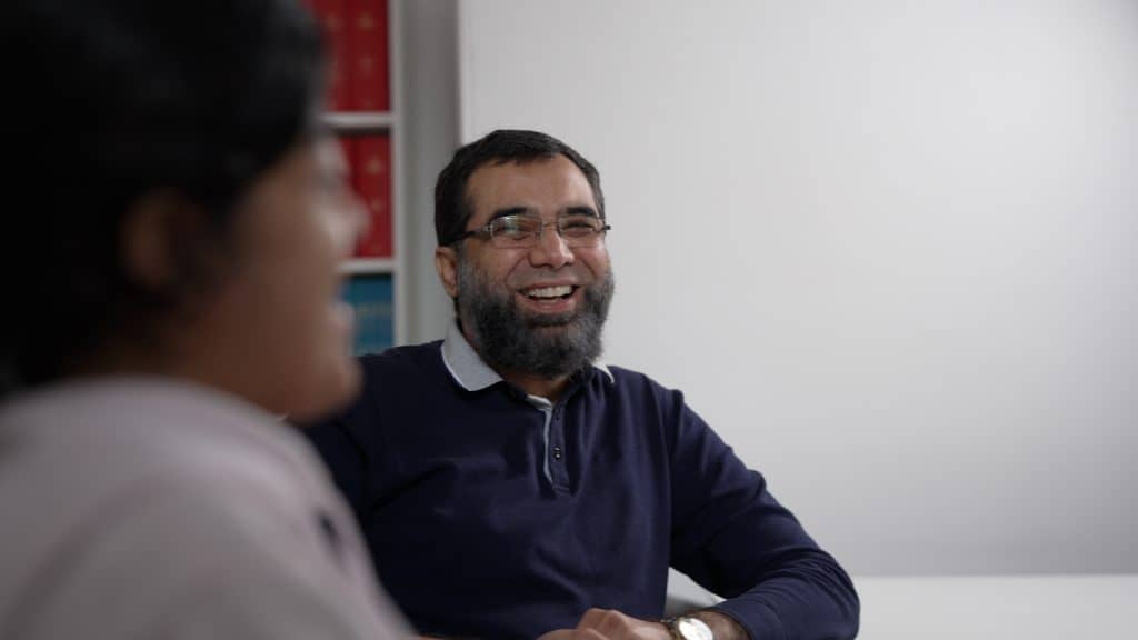 Smiling man in a navy sweater in a clinical setting with a woman in the foreground.